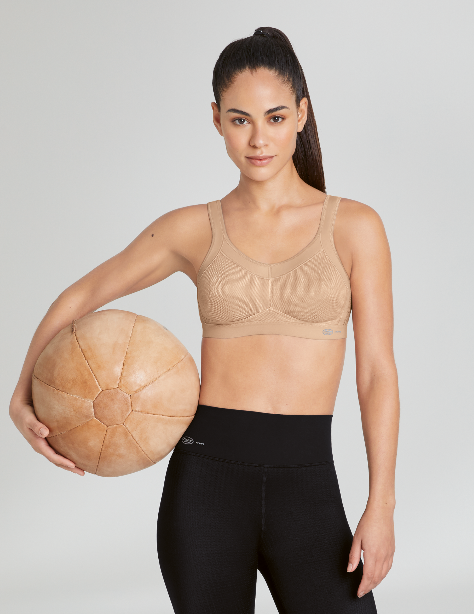 Woman holding a medicine ball wearing a beige sports bra and black leggings on a plain background