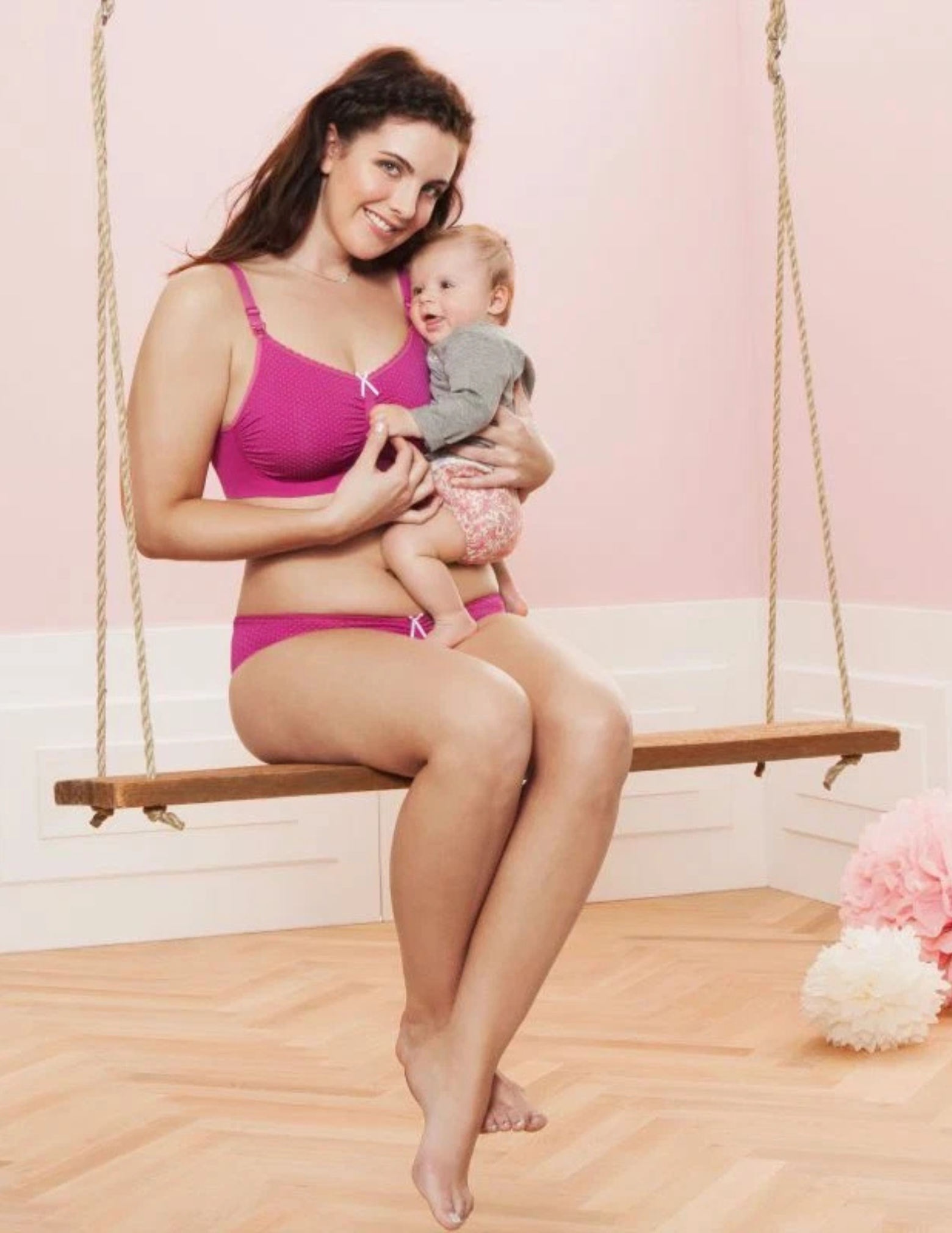 Woman in pink nursing bra and underwear holding a baby on a swing with a pink background